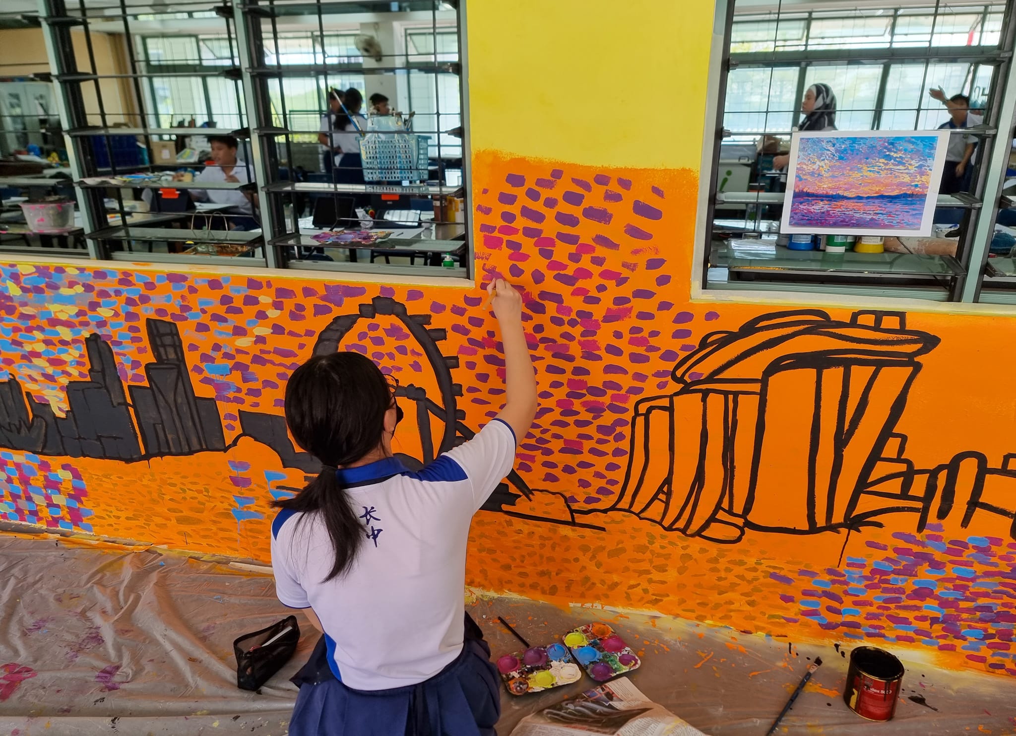 A student in school uniform paints a large orange mural of a city skyline with colorful dabs of paint.